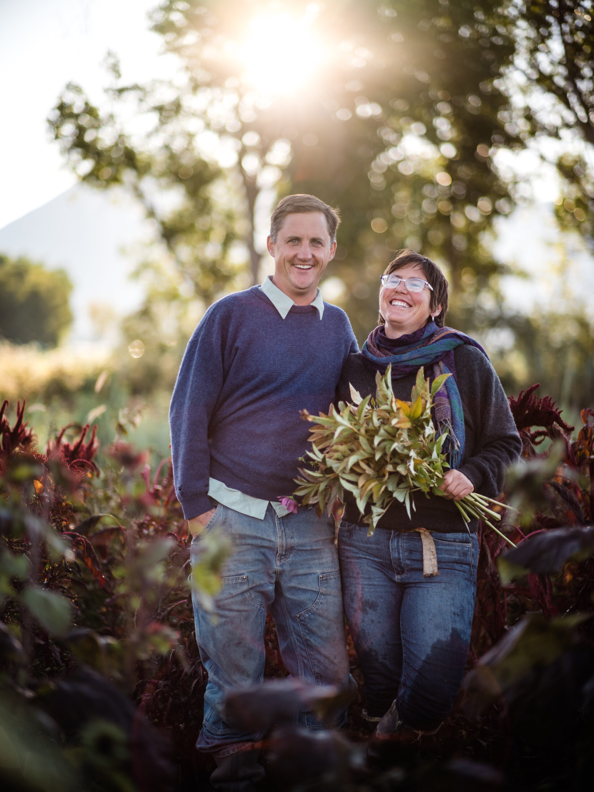 Smiling couple standing in a lush garden, holding a bouquet of fresh herbs, with sunlight filtering through trees in the background. The scene captures the joy of farming and the beauty of nature.