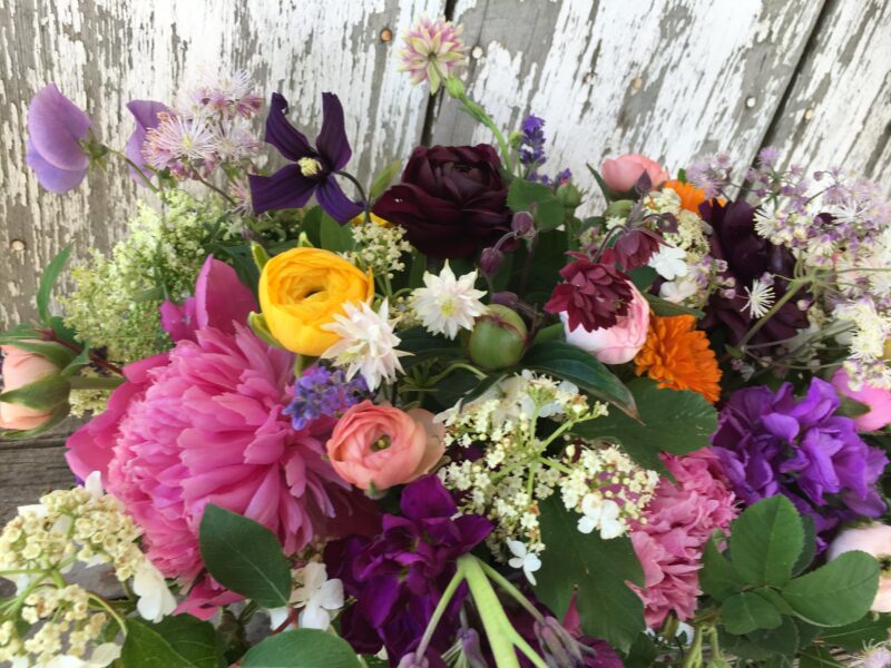 Vibrant bouquet of assorted flowers including peonies, ranunculus, and purple clematis, arranged against a rustic wooden background, showcasing a variety of colors and textures. Ideal for floral decoration or gardening inspiration.