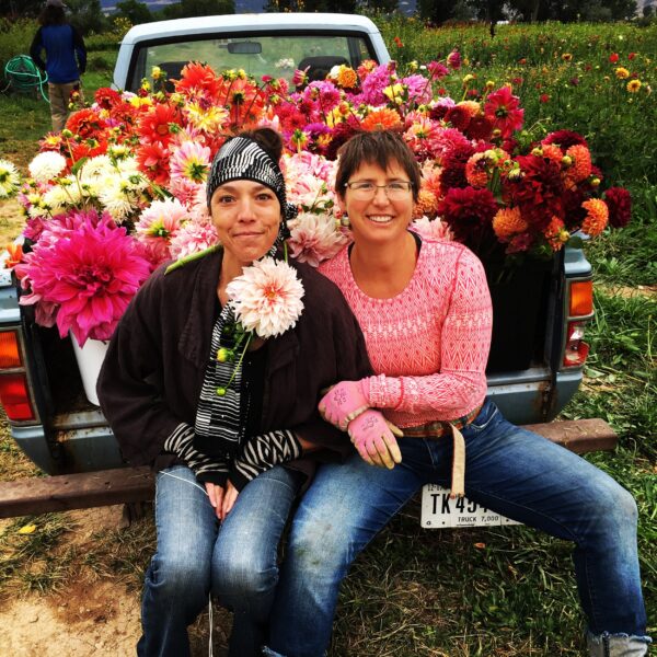 Two women sitting on the tailgate of a truck filled with vibrant flowers, smiling and dressed in casual work attire. The background features a lush garden with colorful blooms, showcasing the beauty of flower farming.