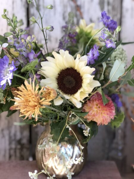 Colorful floral arrangement featuring sunflowers, purple and orange blooms, and greenery in a decorative gold vase, set against a rustic wooden backdrop. Ideal for home decor or as a gift for special occasions.