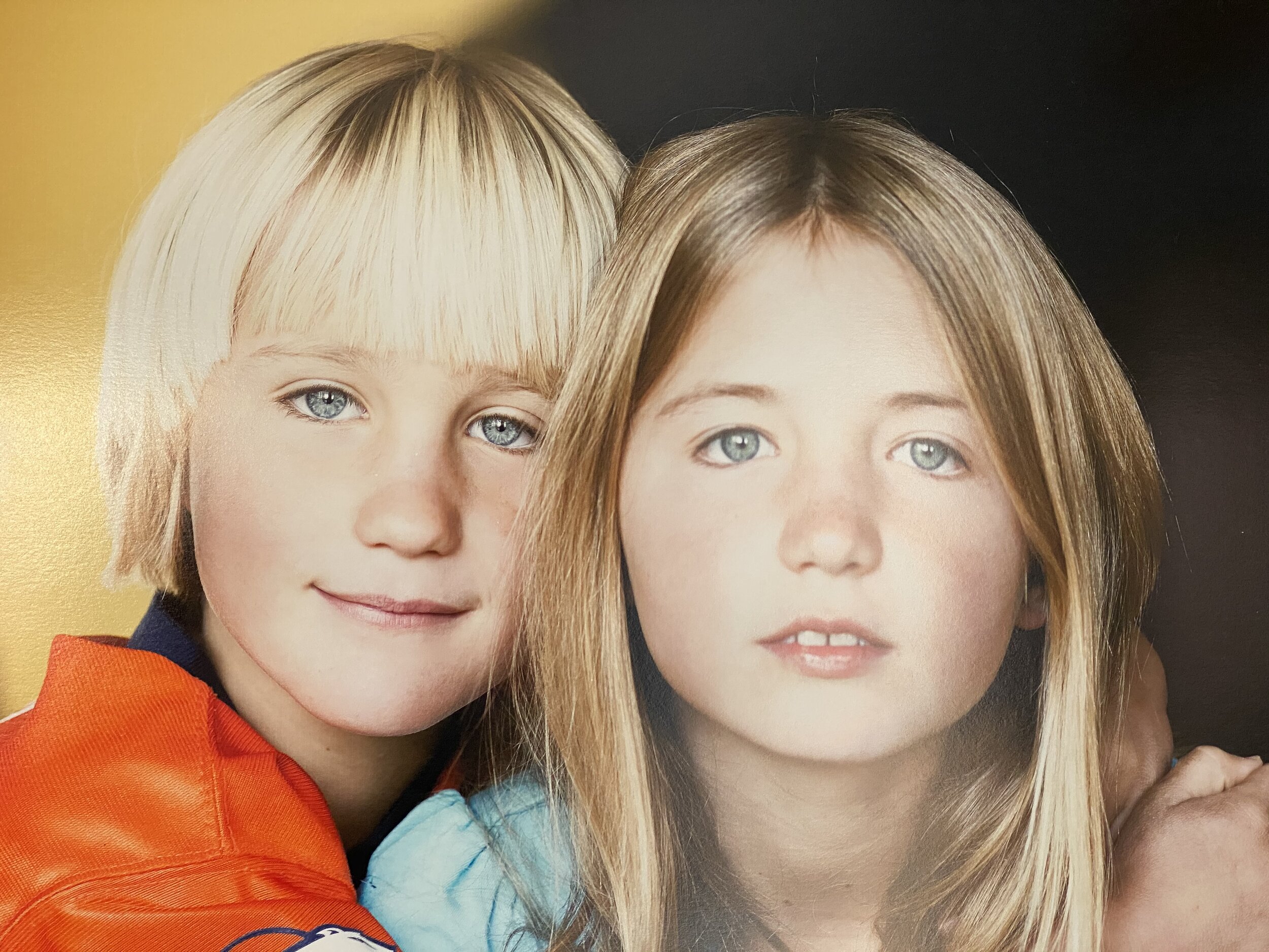Two children with striking blue eyes, one with short blonde hair and the other with long, straight brown hair, pose closely together against a warm, golden background. The child on the left wears an orange sports jacket, while the other wears a light blue top, both smiling gently.